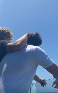 two people sitting on the back of a boat looking out at the water and one person is holding his arm around the other man's shoulder