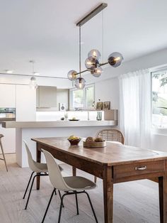 a dining room table with chairs and lights hanging from it's ceiling in front of an open kitchen