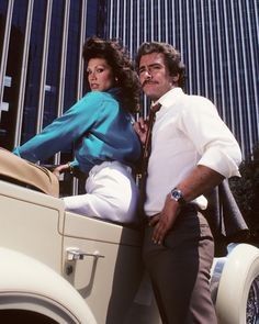 a man and woman leaning on the back of an old car in front of a tall building