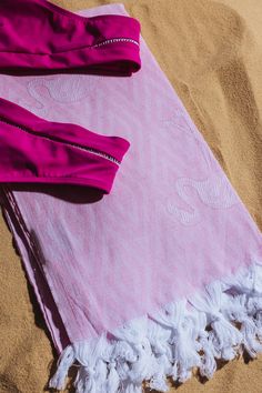 a pink and white towel laying on top of a sand covered beach next to a pair of sunglasses