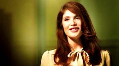 a woman with long brown hair wearing a yellow blouse and smiling at the camera while standing in front of a green wall