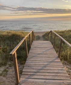 a wooden walkway leading to the beach with grass and water in the background at sunset