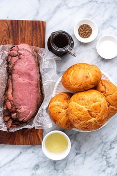 roast beef and bread on a cutting board with dipping sauces next to it, surrounded by other ingredients