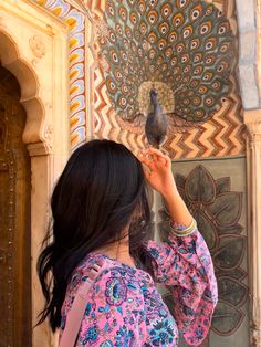 a woman holding a bird in front of a colorful wall with an intricately painted peacock on it