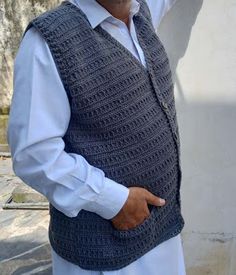 an older man wearing a vest and holding his hand up in the air while standing next to a white wall