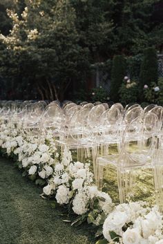 rows of clear chairs with white flowers on the grass in front of trees and bushes