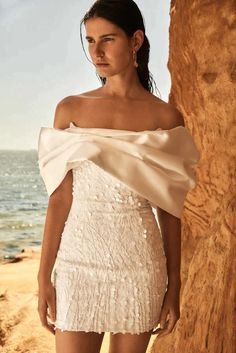 a woman in a short white dress standing on the beach with her arms behind her head