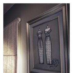 a framed drawing of two dresses hanging on a wall next to a window with sheer drapes