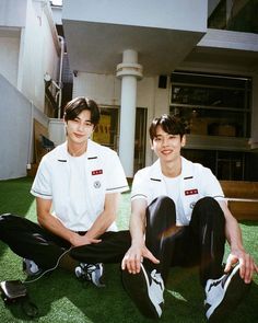 two young men sitting on the grass in front of a building