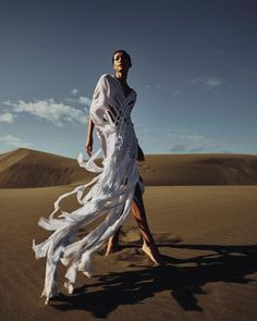 a woman is walking in the desert wearing a white dress with flowing fabric on it