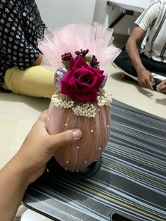 a hand holding a vase with flowers in it on a table next to other people