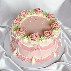 a three tiered cake with pink frosting and roses on the top is sitting on a white table cloth