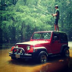 a man standing on top of a red jeep in the middle of a flooded forest