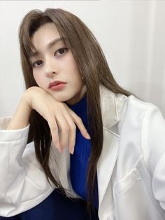 a woman with long brown hair wearing a white jacket and blue dress sitting in front of a white wall