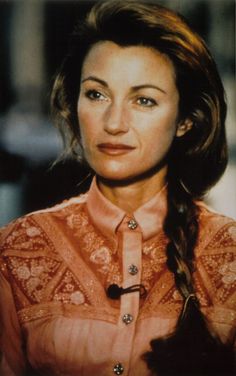 a woman with long hair wearing an orange shirt and braided bangs, looking directly into the camera