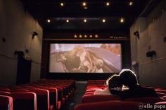 two people are sitting in an auditorium watching a movie on the big screen with red chairs