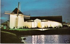 a windmill is lit up in front of a white building with a water reflection on the ground