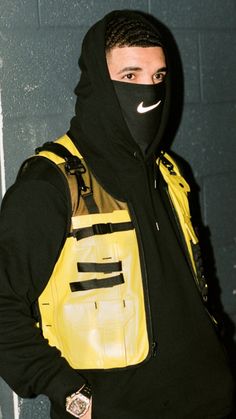 a man wearing a yellow vest and black hoodie standing in front of a wall