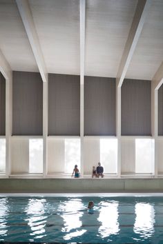 two people sitting on the edge of a swimming pool in front of large windows with vertical blinds