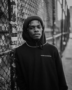 a young man leaning against a fence wearing a hoodie and looking at the camera