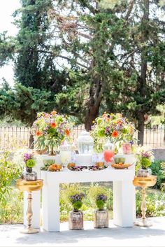 an outdoor table with potted plants and flowers on it in front of some trees