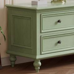 a green dresser with two drawers and a clock on it's top, next to a potted plant