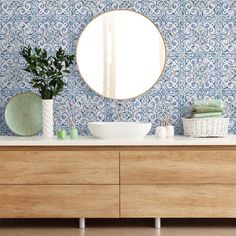 a bathroom with blue and white wallpaper, a round mirror above the sink and wooden cabinets