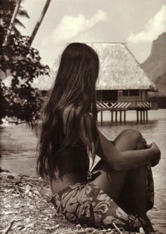 a woman is sitting on the beach looking out at the water and huts in the background