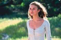 a beautiful young woman standing in front of a lush green field wearing a white top