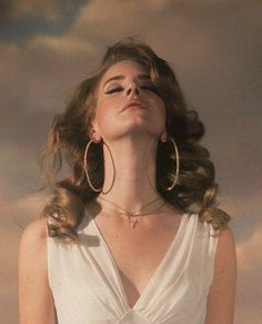 a woman with her eyes closed wearing large gold hoop earrings and a white dress in front of a cloudy sky