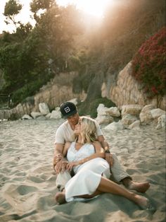 a man and woman sitting in the sand on a beach with their arms around each other