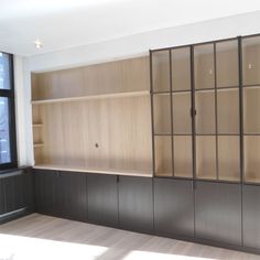 an empty room with wooden shelves and black cupboards on the wall, in front of a large window