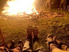 two people sitting in front of a campfire with their feet up on the ground