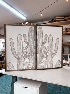 two framed pictures with cactus designs on them sitting on a table in front of bookshelves