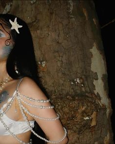 a woman with makeup and pearls on her chest standing next to a tree wearing a white bra