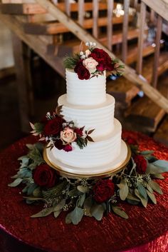 a three tiered white cake with red flowers and greenery on the table next to stairs