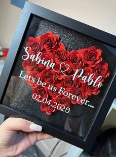 a person holding up a black frame with red roses in the shape of a heart