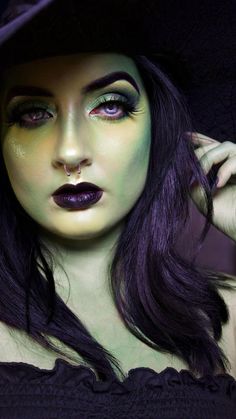 a woman with dark hair and green makeup wearing a black witches hat, holding her hand to her ear