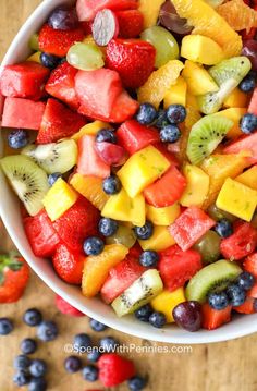 a white bowl filled with fruit salad on top of a wooden table next to blueberries, kiwis and strawberries