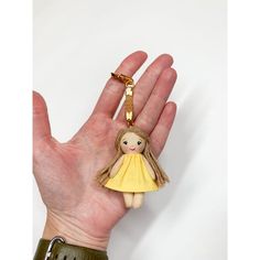 a hand holding a small doll with a yellow dress on it's back and a gold key chain attached to it