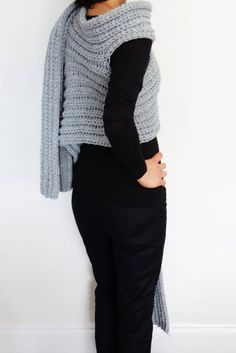 a woman wearing a gray knitted shawl and black pants standing in front of a white wall