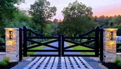 a wooden gate with stone pillars leading to a grassy field at sunset or dawn in the background