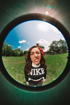 a girl is seen through the lens of a mirror in a grassy area with trees and blue sky