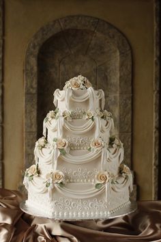 a three tiered white wedding cake with flowers on it's side and an archway in the background