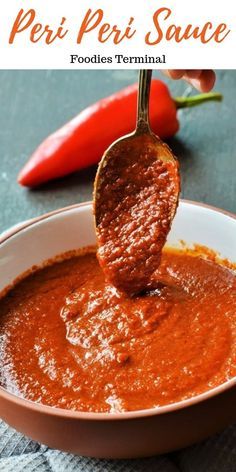 a spoon full of red sauce being held up by a person's hand with peppers in the background