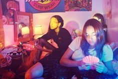 two people sitting on a bed in a room with neon lights and posters above them