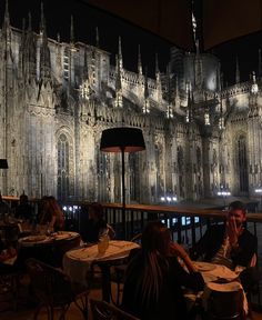 people sitting at tables in front of a large building with lights on it's side