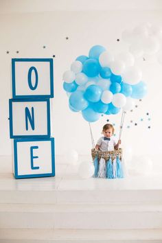 a baby boy sitting in a basket with blue and white balloons on top of it