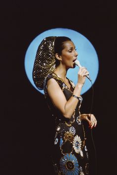 a woman in an egyptian dress singing into a microphone with a blue circle behind her