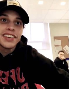 a young man wearing a black hoodie and cap smiles at the camera while standing in an office